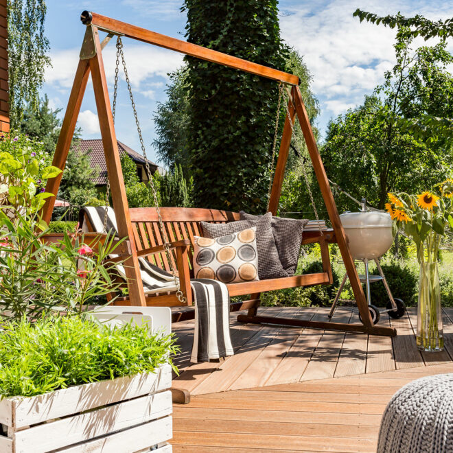 A garden swing seat on decking. It is surrounded by flowers.